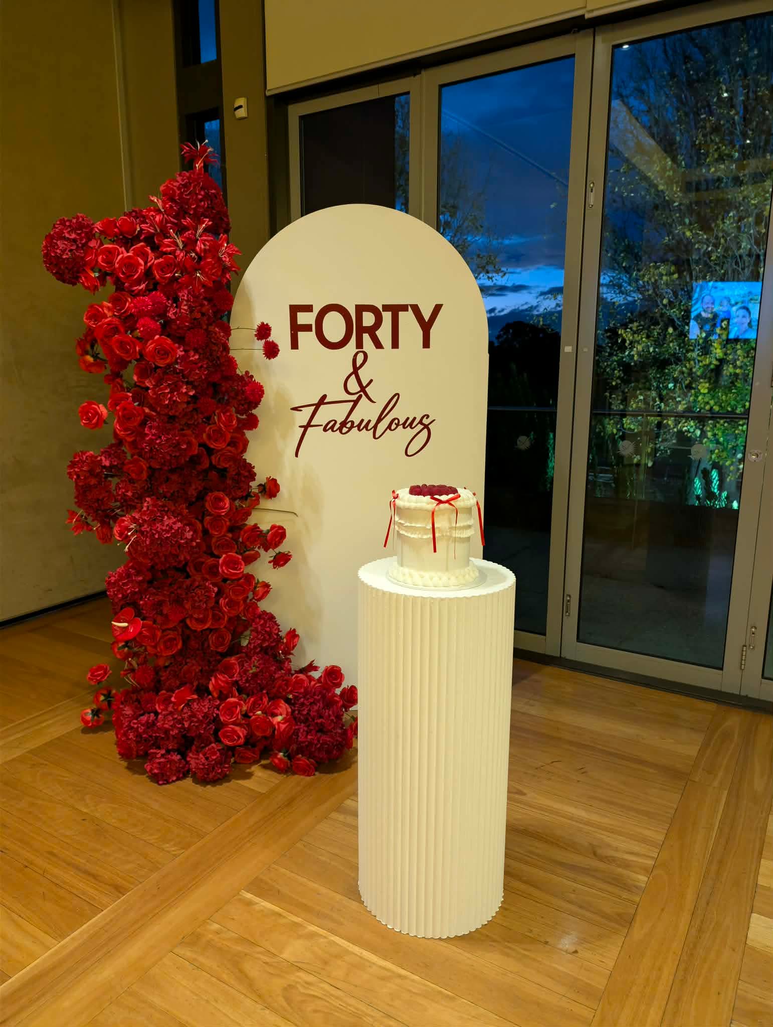 Forty and Fabulous 40th birthday backdrop with red rose floral arch and cake on white ribbed plinth