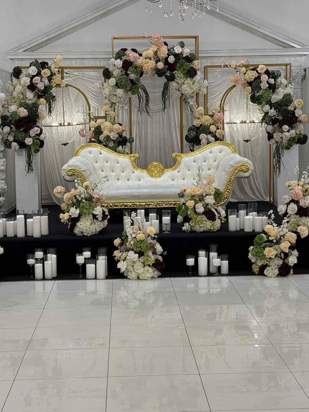 Wedding stage with white and gold chaise, gold frame arches, crystal chandeliers and peach and burgundy florals